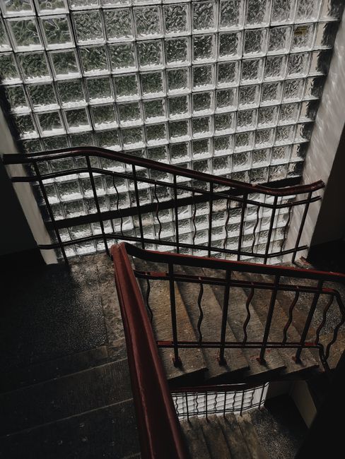 A view of a narrow, indoor staircase within a building, showing dark wooden handrails and metal balustrades with vertical bars. The staircase appears to be part of a residential property in Osterley, with the steps made of dark material, possibly concrete or stone. The walls around the staircase are partially visible, and there is a large glass block window at the corner, allowing diffuse natural light to illuminate the area. The image captures a moment during a home relocation or furniture transport process, typical of house removals in the Osterley area, as handled by Man with Van Osterley, with the staircase being a challenging element for moving large items between floors.