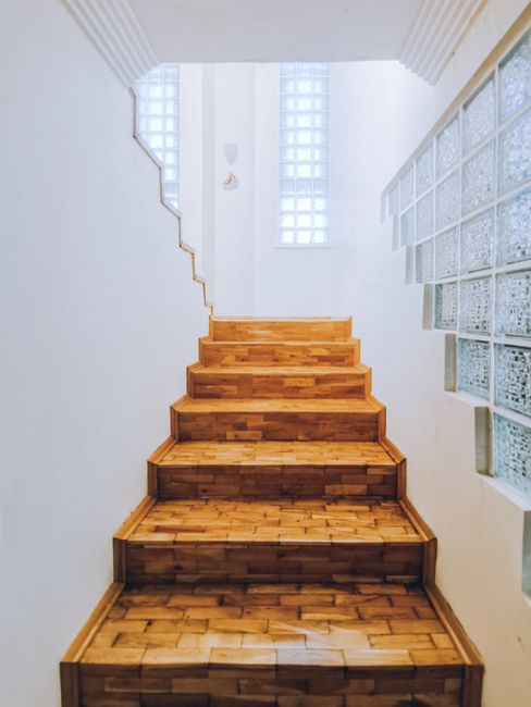 A set of wooden stairs with five steps leading upward inside a property, with a white wall on the left side and a glass block wall on the right side. The stairs are made of polished wood, showing a natural grain and a warm tone, and are situated in a narrow corridor. At the top of the stairs, there is a small window composed of glass blocks, allowing diffused light into the space. The surrounding environment appears bright and clean, with minimal decoration. Nearby, a glass brick wall separates the staircase area from an adjacent room or hallway, and the lighting is natural, casting soft shadows. This interior stairway could be involved in home relocation or moving procedures, where professionals like Man with Van Osterley might assist with furniture transport and packing within flats. The image depicts the challenging nature of blocked stairs in Osterley flats and demonstrates potential solutions used during moving services, focusing on the staircase's structure and environment for effective logistics.