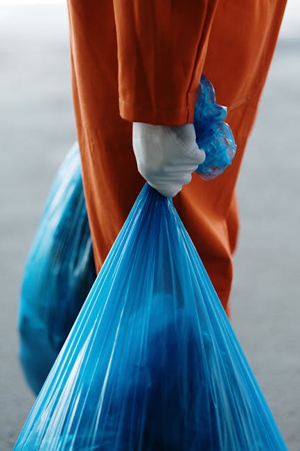 A person wearing white gloves and an orange protective suit is holding a large blue plastic garbage bag filled with waste material. The individual is standing outdoors on a paved surface, which appears to be part of a residential or commercial area, with an empty or unobtrusive background. This scene reflects waste collection or disposal activities, often associated with clearing items during house removals or home decluttering. The image highlights the importance of proper waste management in the context of moving or furniture transport, as handled by companies like Man with Van Osterley, who assist with house removals, including bulky waste disposal. The focus on the bag's volume and the person's grip underscores the physical effort involved in removing waste, an essential step in the packing and moving process to prepare a property for relocation or renovation.