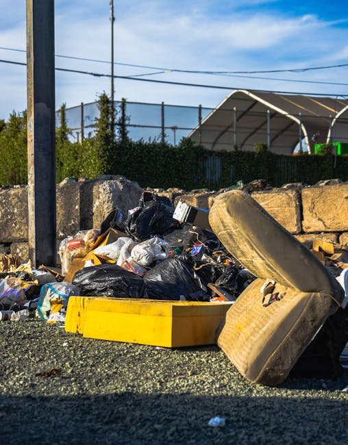 An outdoor scene showing a pile of bulky waste and debris, including several black plastic rubbish bags, a large yellow plastic container, and a worn-out upholstered armchair with a cushion, all placed on a paved surface next to a stone wall. In the background, there are metal fences, green bushes, utility poles with overhead power lines, and a partly cloudy sky. The image appears to be taken during daylight hours. The scene relates to disposal and clearance of waste before or during a house relocation process, as managed by Man with Van Osterley, a professional removals company specializing in house removals and waste removal services.