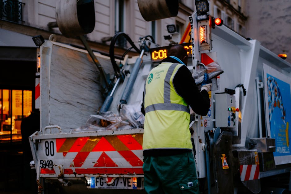 A person wearing white gloves and an orange protective suit is holding a large blue plastic garbage bag filled with waste material. The individual is standing outdoors on a paved surface, which appears to be part of a residential or commercial area, with an empty or unobtrusive background. This scene reflects waste collection or disposal activities, often associated with clearing items during house removals or home decluttering. The image highlights the importance of proper waste management in the context of moving or furniture transport, as handled by companies like Man with Van Osterley, who assist with house removals, including bulky waste disposal. The focus on the bag's volume and the person's grip underscores the physical effort involved in removing waste, an essential step in the packing and moving process to prepare a property for relocation or renovation.