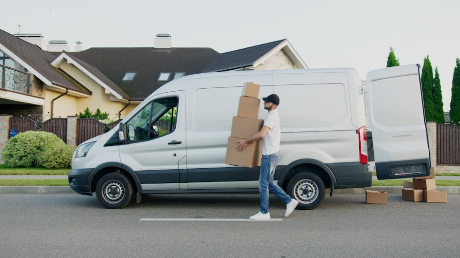 A man wearing a white t-shirt and jeans is standing on the sidewalk next to an open white moving van, which is parked in front of a suburban house with a pitched roof and a tall green shrub. The man i