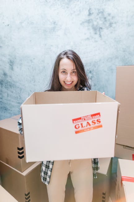 A woman with long brown hair, smiling and standing indoors against a light blue textured wall, holding an open cardboard box labelled 'Fragile Glass' in red text. The box is empty and appears to be pr