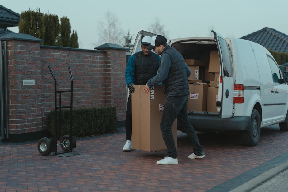 Two men dressed in dark and casual clothing are engaged in a home relocation process, standing outside a residential property on a paved driveway. They are lifting and inspecting a large cardboard box, part of the packing and moving activities. Nearby, a hand truck with black handles and wheels is positioned on the ground, ready for use. Behind them, an open white van with its rear doors swung open reveals multiple packed cardboard boxes of various sizes stacked inside, indicating an organized furniture transport and packing process. The garage and brick wall of the property are visible in the background, along with some greenery and trees under an overcast sky. This scene reflects the logistics involved in professional removals, as provided by Man with Van Osterley, supporting efficient moving and relocating services in the Osterley area as outlined in the Hounslow Council permit guide for Osterley moving vans.