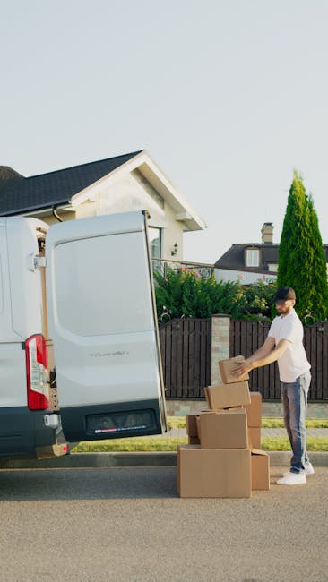 A man, dressed in a white t-shirt and jeans, is standing on the street beside an open white moving van used for home relocation. He is placing a cardboard box onto a stack of similar boxes, which are arranged on the pavement next to the vehicle. The van's rear door is open, revealing a spacious cargo area with some packaging materials, likely for furniture and household items. The background features residential houses, a wooden fence, and tall, green conifer trees under a clear sky. The scene captures the loading process typical of furniture transport and packing during a house removal, with the man preparing goods for safe transport with the assistance of the moving vehicle, representing professional removal services by Man with Van Osterley.
