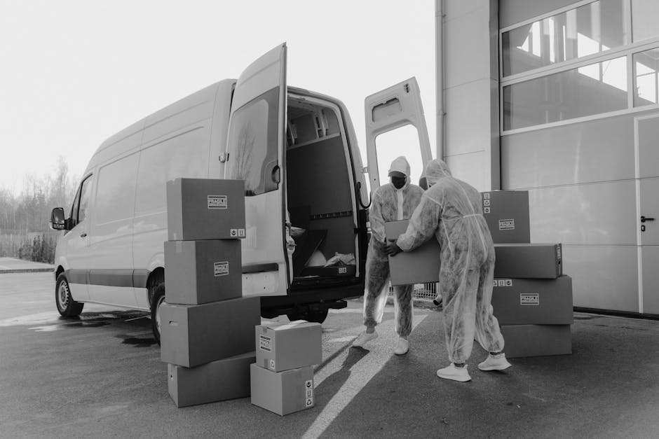 Two men dressed in dark and casual clothing are engaged in a home relocation process, standing outside a residential property on a paved driveway. They are lifting and inspecting a large cardboard box, part of the packing and moving activities. Nearby, a hand truck with black handles and wheels is positioned on the ground, ready for use. Behind them, an open white van with its rear doors swung open reveals multiple packed cardboard boxes of various sizes stacked inside, indicating an organized furniture transport and packing process. The garage and brick wall of the property are visible in the background, along with some greenery and trees under an overcast sky. This scene reflects the logistics involved in professional removals, as provided by Man with Van Osterley, supporting efficient moving and relocating services in the Osterley area as outlined in the Hounslow Council permit guide for Osterley moving vans.