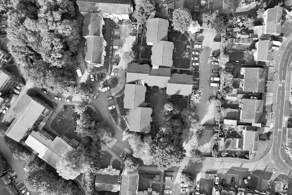 Aerial black and white photograph showing a residential area with multiple detached and semi-detached houses, some with driveways filled with parked cars. The houses have pitched roofs and are surrounded by trees and greenery. A narrow pathway runs between the houses, leading to the main street which curves around the estate. In the foreground, a private driveway with cars parked along its sides leads to the rear of the houses, and a small garden area is visible with trees and bushes. The image captures a section of a suburban neighbourhood, likely during daytime with natural light illuminating the scene. The image is used in the context of house removals, showcasing the environment where a professional service such as [COMPANY_NAME] might operate, coordinating moving logistics around narrow access points and planning home relocations within this residential community.