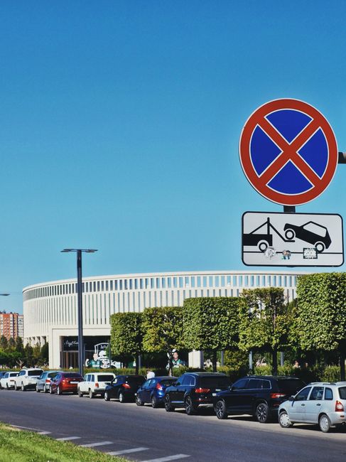A row of parked cars along a street in front of a modern, curved building with a white exterior and large windows, under a clear blue sky. In the foreground, there is a prominent no parking sign with a blue circle crossed by red lines, mounted on a pole, accompanied by a smaller black and white sign indicating towing enforcement. The scene suggests an urban environment suitable for house removals or furniture transport activities, with the location possibly requiring parking suspensions or compliance with local regulations. The presence of parked vehicles, signage, and the building's architecture reflect a typical setting for a home relocation service such as Man with Van Osterley, involving loading and unloading operations within a city context.