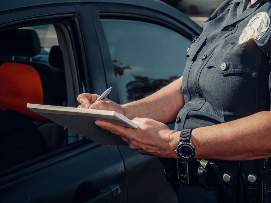 A police officer wearing a dark grey uniform with a badge on the chest and a black wristwatch on their left wrist is standing next to a black vehicle with tinted windows. The officer is holding a clipboard with a notepad attached and is writing on it with a silver pen. The vehicle is parked outdoors, and the interior is partially visible through the window, showing a black and orange seat cover inside. The background includes some trees and a cloudy sky. This scene reflects official parking enforcement and compliance checks, which may be relevant in the context of house removals and moving logistics managed by Man with Van Osterley, ensuring compliance with parking regulations during home relocation or furniture transport.