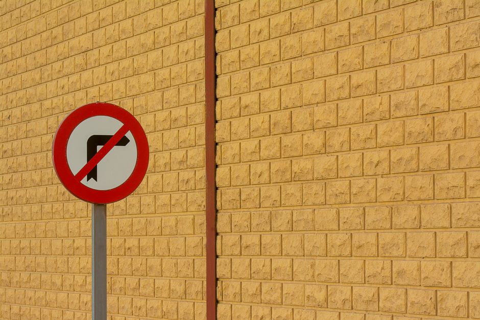 A round traffic sign with a red border and a white background, displaying a black arrow pointing left with a red slash through it, indicating no left turn. The sign is mounted on a grey metal pole positioned in front of a brick wall painted in yellow-beige tones. The wall features evenly spaced rectangular bricks with mortar joints, and is illuminated by natural daylight. Part of a pavement surface is visible at the base of the sign, with a small gap between the wall and ground. The scene is neutral and professional, illustrating street rules relevant to home relocation, furniture transport, or moving logistics managed by Man with Van Osterley, particularly concerning pavement and double-yellow line regulations for Osterley removals.