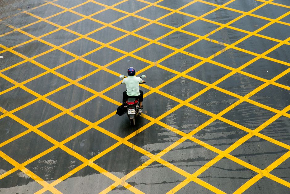 A person riding a motorcycle across a large paved area, which is marked with a grid of bright yellow crisscrossing lines. The rider is wearing a helmet and casual clothing, with their back facing the camera. The pavement appears wet, with reflections visible on the surface, and is surrounded by an organized, regulated environment. This scene likely takes place in a designated parking or loading zone, where pavement and double-yellow line rules govern parking and waiting regulations. The image is associated with house removals and transportation services by Man with Van Osterley, highlighting the importance of adhering to parking regulations during the home relocation or furniture transport process, especially in urban areas with specific pavement markings and yellow lines.