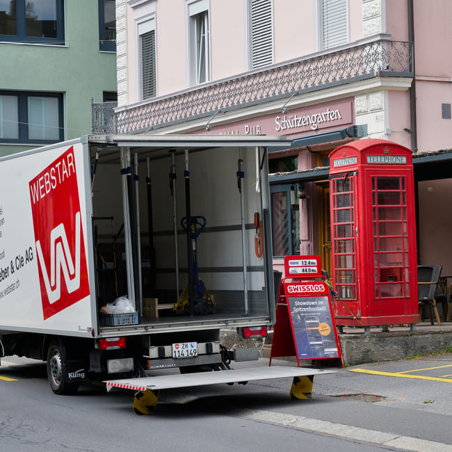 A white moving van with open side doors is parked on a street in front of a building with a pink exterior, a balcony with decorative railings, and a sign for a pub and beer garden. Inside the van, various packing materials such as cardboard boxes, plastic wrap, and lifting straps are visible, along with hand trucks and moving equipment. The van is equipped with yellow and black wheel stabilizers to ensure stability during the loading process. Nearby, a traditional red telephone booth and a black chair are situated on the pavement, indicating an urban residential area. The scene captures the preparation phase of home relocation, with furniture and boxes ready for transport, aligning with services offered by Man with Van Osterley for efficient furniture transport and packing and moving tasks in the Osterley locality.