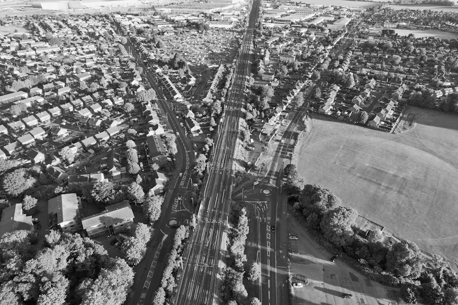 A black and white aerial photograph showing a suburban residential area with numerous detached and semi-detached houses, each with individual driveways and gardens, adjacent to a large open field on the right side. The image captures a wide main road with multiple lanes and tram tracks running through the centre, intersected by smaller roads and roundabouts. Trees line the streets and the open field, creating a natural boundary. In the foreground, a section of a building or fencing is visible, indicating the location of a property or a closed entrance. The overall scene depicts a typical home neighbourhood with organized streets, green spaces, and local infrastructure, suitable for illustrating house removals, packing and moving, or furniture transport processes as part of a house relocation in Osterley, with Man with Van Osterley potentially involved in logistical support for these moves.