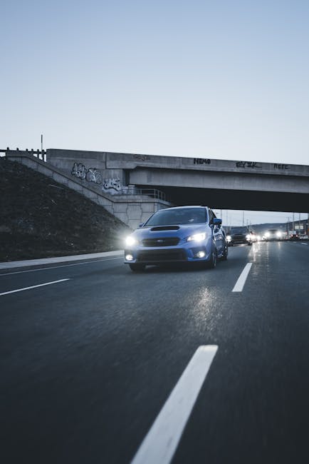 A silver vehicle is driving on a multi-lane urban road during dusk, with headlights on and other vehicles visible in the background. The road has clear white lane markings, and an overpass with graffiti on the concrete supports is situated above the traffic. The scene is well-lit by natural fading daylight, and the surrounding environment includes a sloped embankment on one side. This setting depicts a typical urban transport environment, relevant for illustrating house removal logistics, vehicle transport, and the transit phase of home relocation services offered by Man with Van Osterley, supporting efficient furniture transport and packing and moving processes.
