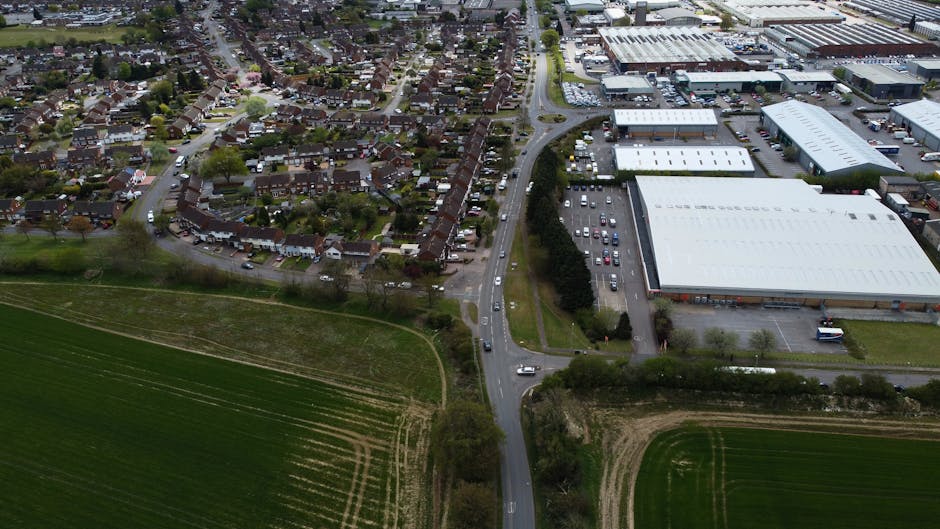 A black and white aerial photograph showing a suburban residential area with numerous detached and semi-detached houses, each with individual driveways and gardens, adjacent to a large open field on the right side. The image captures a wide main road with multiple lanes and tram tracks running through the centre, intersected by smaller roads and roundabouts. Trees line the streets and the open field, creating a natural boundary. In the foreground, a section of a building or fencing is visible, indicating the location of a property or a closed entrance. The overall scene depicts a typical home neighbourhood with organized streets, green spaces, and local infrastructure, suitable for illustrating house removals, packing and moving, or furniture transport processes as part of a house relocation in Osterley, with Man with Van Osterley potentially involved in logistical support for these moves.