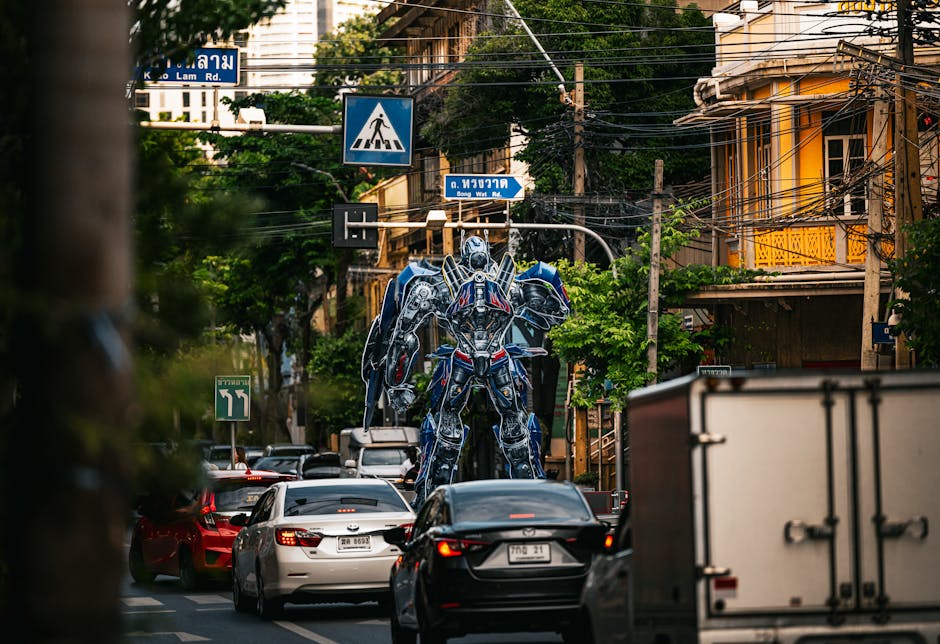 A busy urban street scene with multiple cars waiting in traffic, situated beneath overhead street signs indicating Lampton Road and Song Wat Road. In the background, there is a large, detailed sculpture of a robot made from metallic and plastic materials standing between buildings, which include structures with yellow balconies and orange walls, surrounded by green trees. Utility poles and a network of electrical wires run across the scene. The street appears to be in a city area suited for house removals and moving logistics, with visible vehicles in the foreground and a clear focus on the urban environment where moving services like those offered by Man with Van Osterley could operate during home relocation or furniture transport activities.